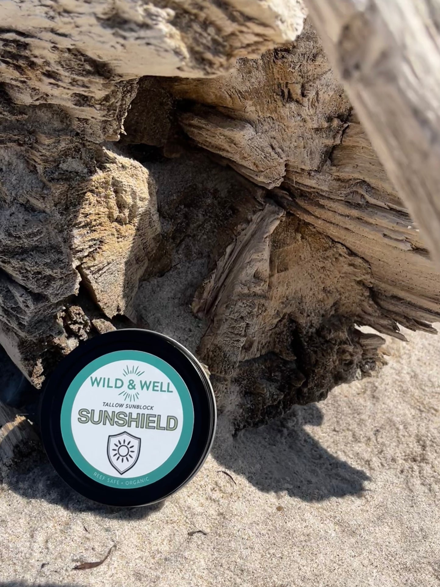 Container of Wild & Well Sunshield on a sandy surface with driftwood