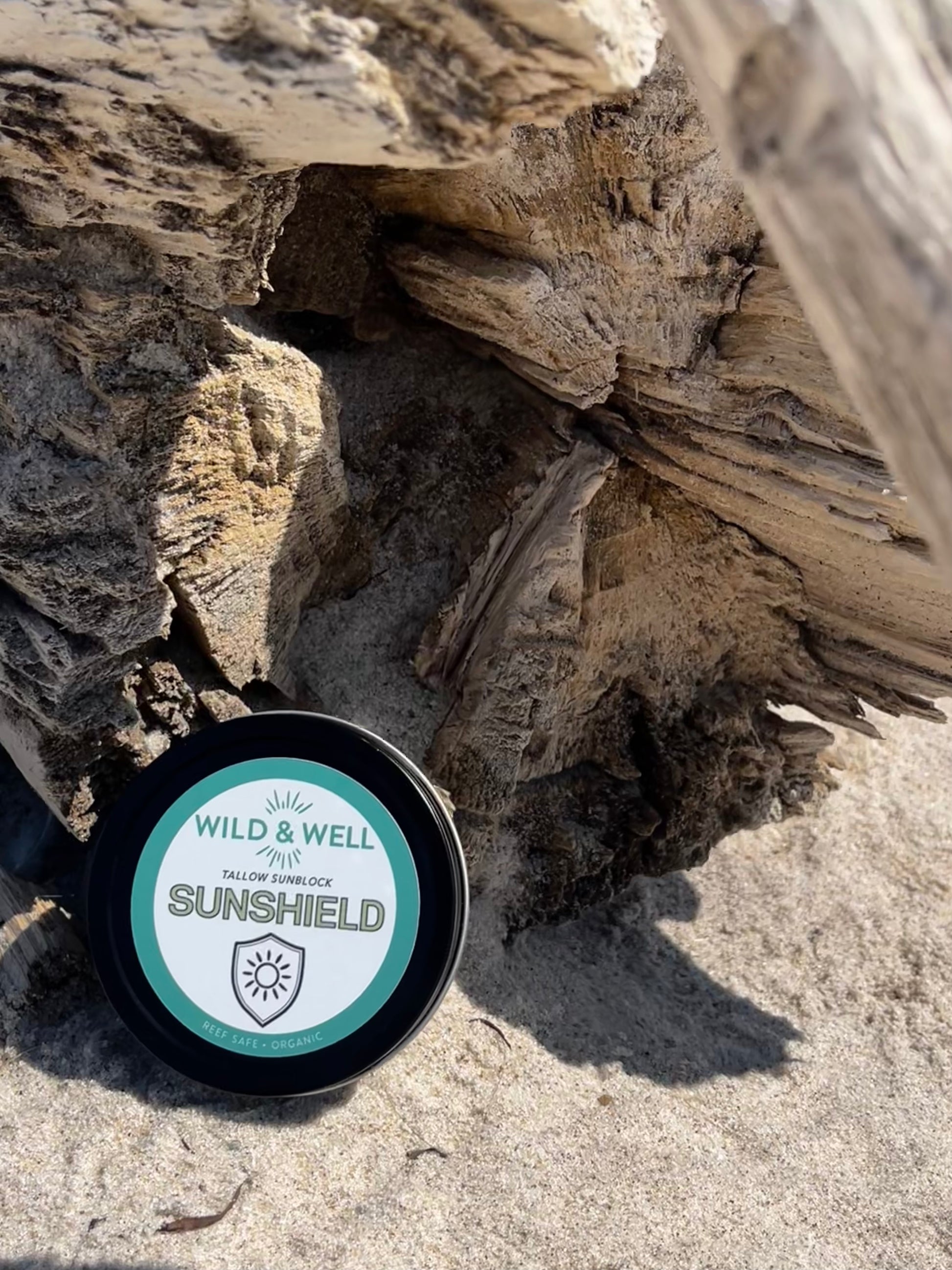 Container of Wild & Well Sunshield on a sandy surface with driftwood